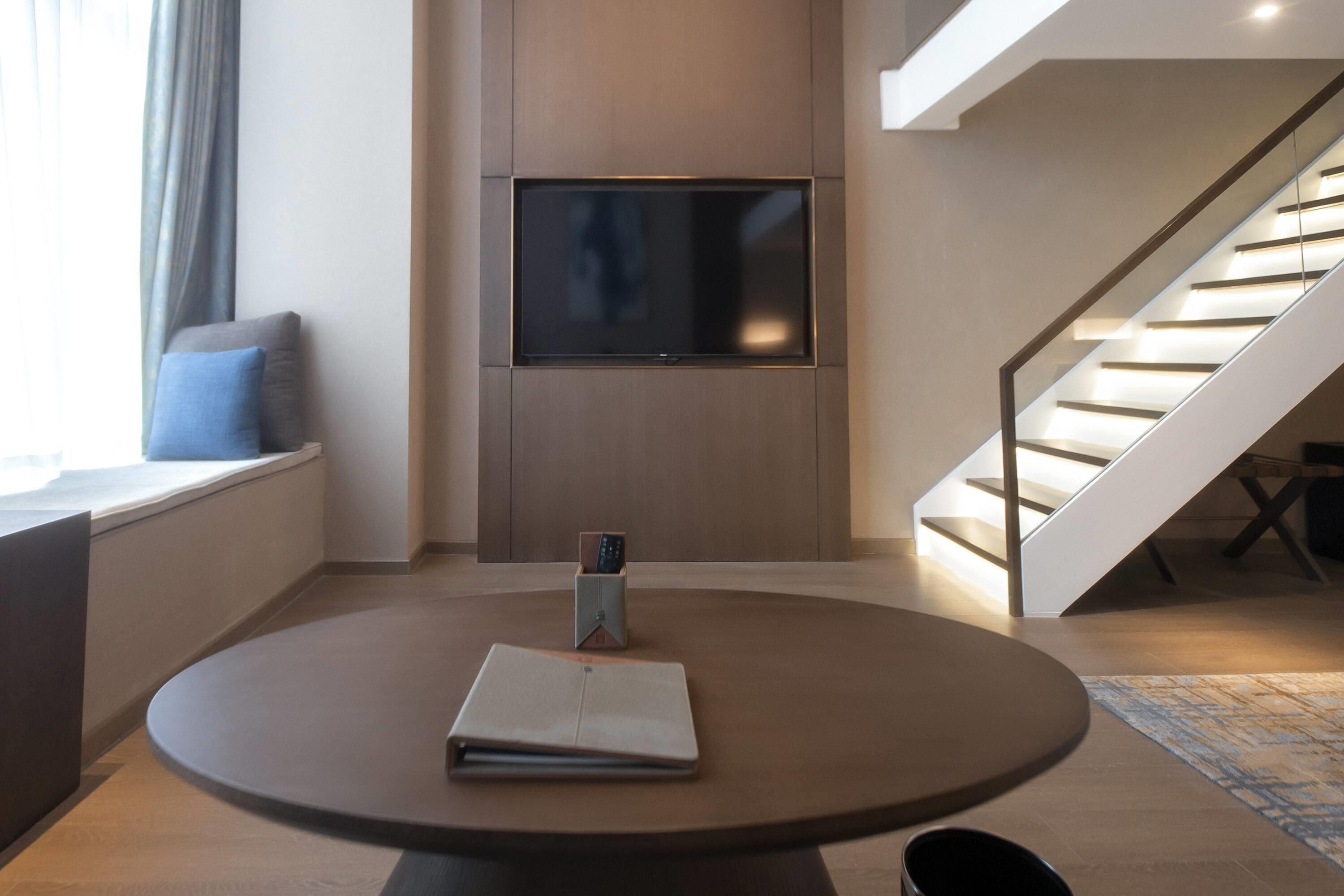 A modern living room with a round table, a flat-screen TV mounted on the wall, and a staircase with illuminated steps.
