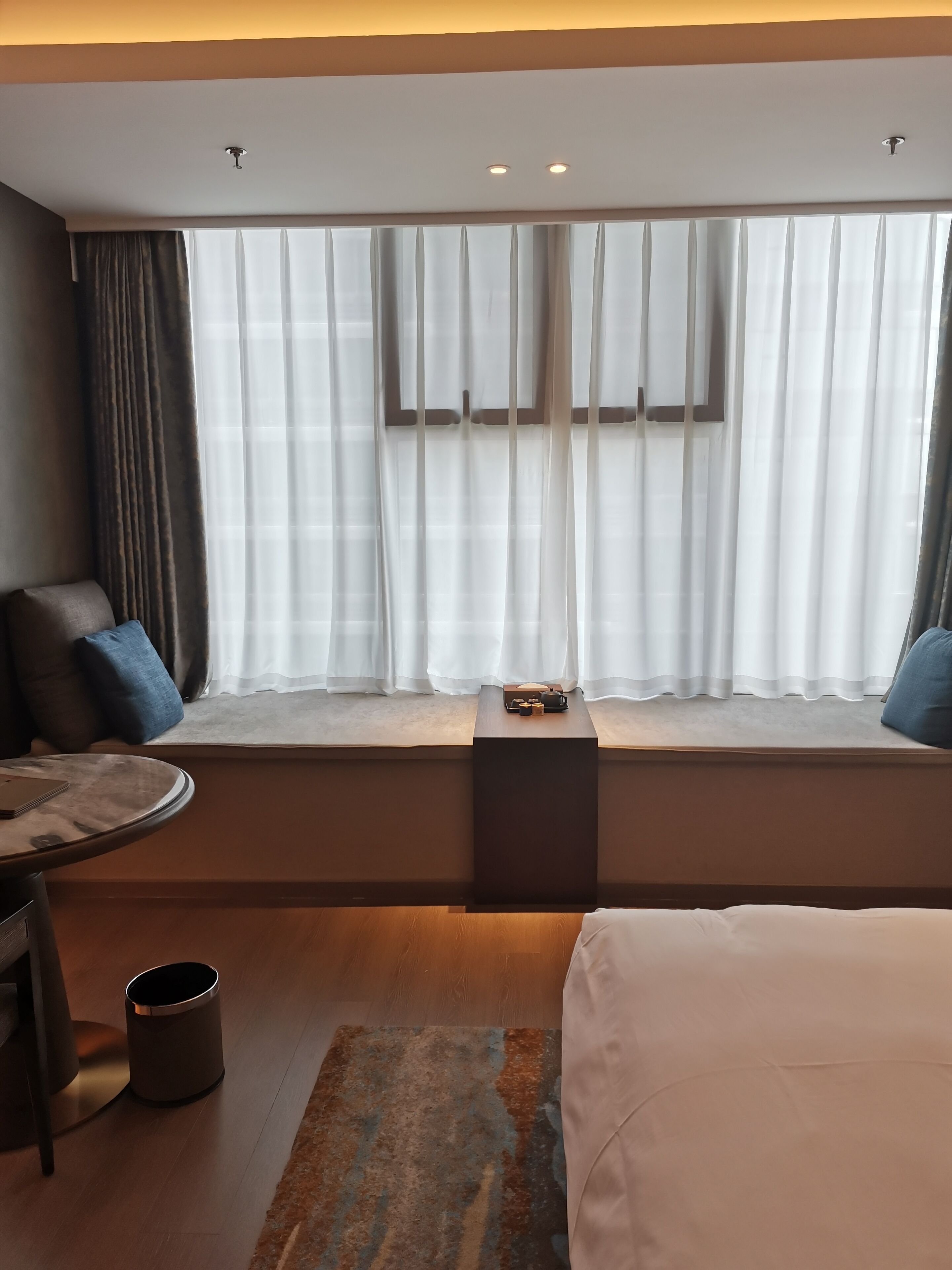 Une chambre d’hôtel avec une grande fenêtre, un lit, une table de chevet, une chaise et une petite table avec un plateau.