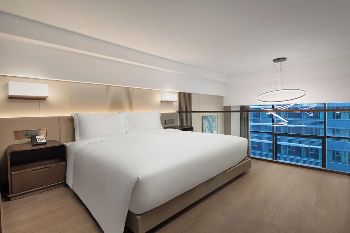 Ett modernt hotellrum med en stor säng, sängbord, en telefon och utsikt över stadsbilden genom stora fönster.