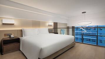 Habitación de hotel moderna con una cama grande, mesitas de noche, teléfono y vistas a un paisaje urbano a través de amplios ventanales.
