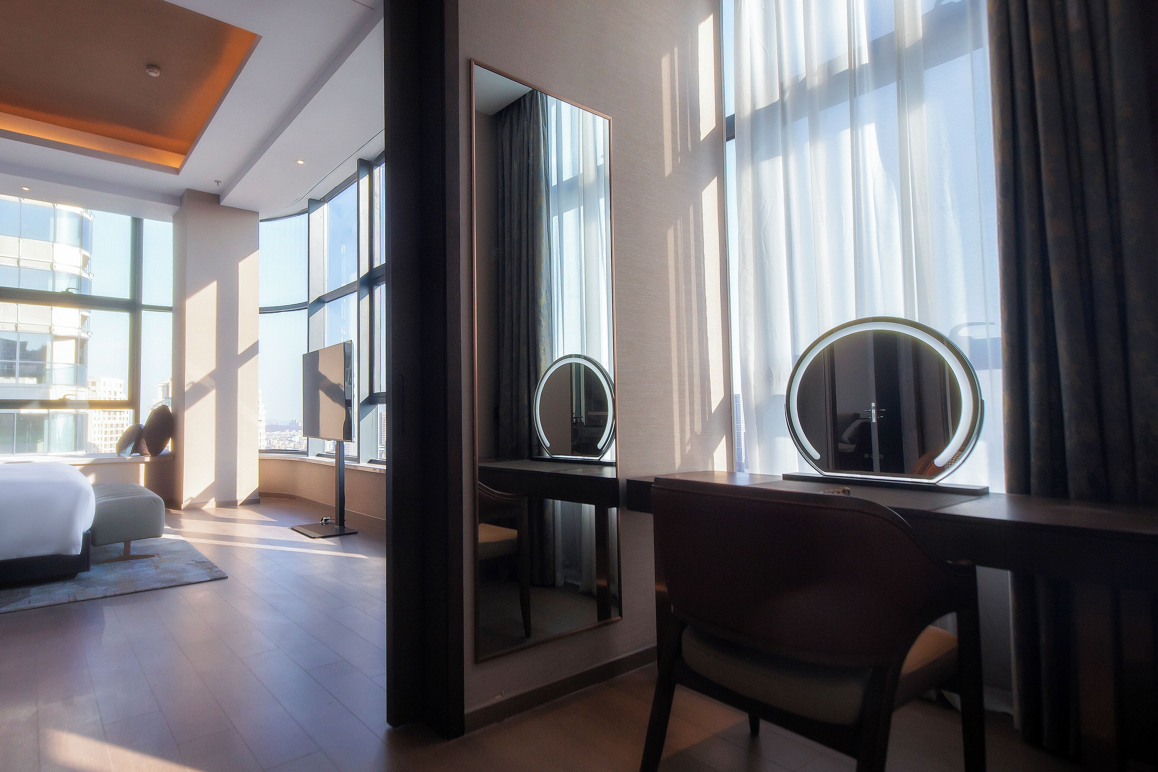 A modern hotel room with a large bed, a desk with a round mirror, and a view of the city through large windows.