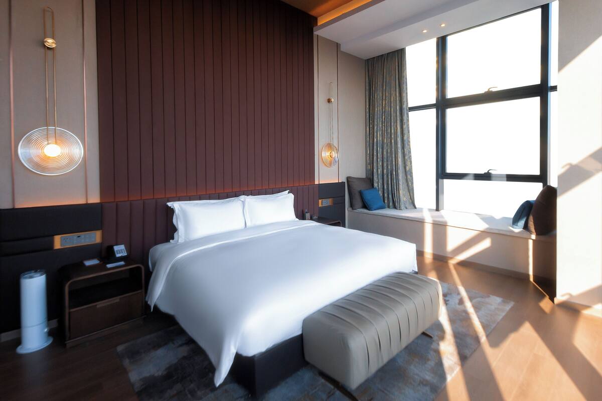 Ett modernt hotellrum med en stor säng, sängbord, ett litet bord, en bänk och ett stort fönster med gardiner.