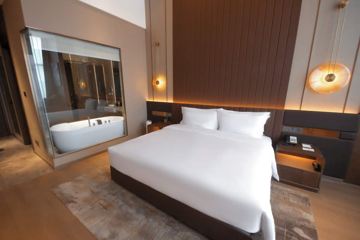 Ett modernt hotellrum med en stor säng, ett badrum med glasväggar och en sänggavel i trä.