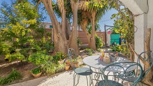 Une terrasse avec une table ronde et des chaises en métal, entourée de plantes en pot et d’arbres.