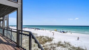 Vue sur la plage depuis une terrasse en bois dotée d’une rambarde, avec une plage de sable et un ciel bleu clair.