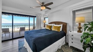 Une chambre avec un grand lit, un ventilateur de plafond, une table de chevet avec une lampe, et une vue sur la plage grâce à de grandes fenêtres.