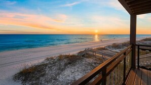Vue sur la plage depuis une terrasse en bois dotée d’une balustrade, avec vue sur l’océan et un coucher de soleil.