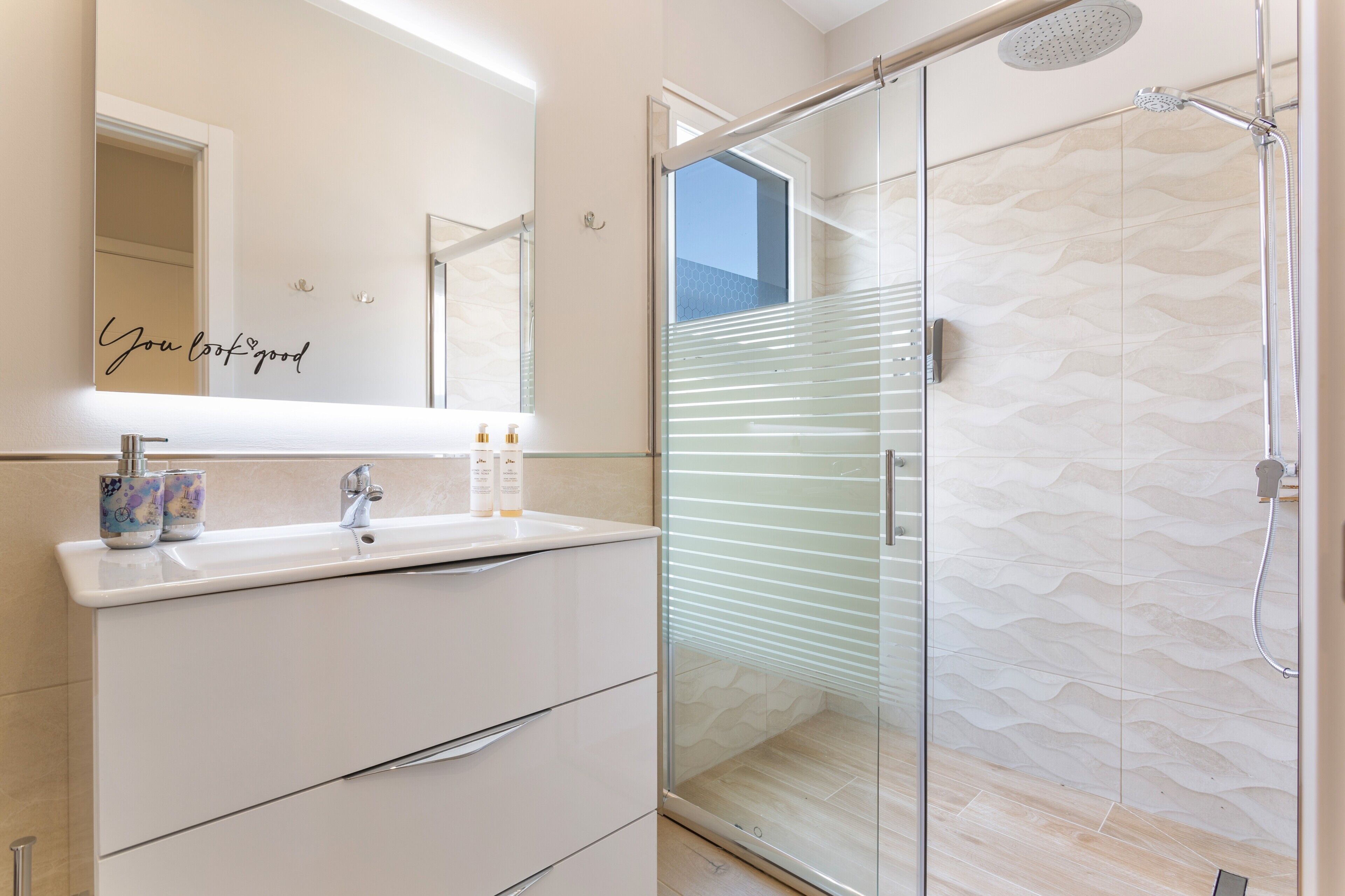 Un baño moderno con cabina de ducha de vidrio, un tocador blanco con espejo y una frase decorativa en el espejo.