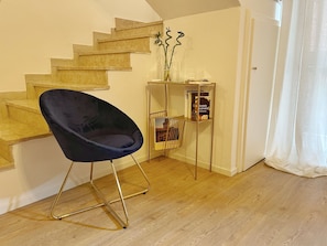 Una habitación con una silla azul oscuro, una escalera de madera, un estante metálico con libros y una puerta blanca.