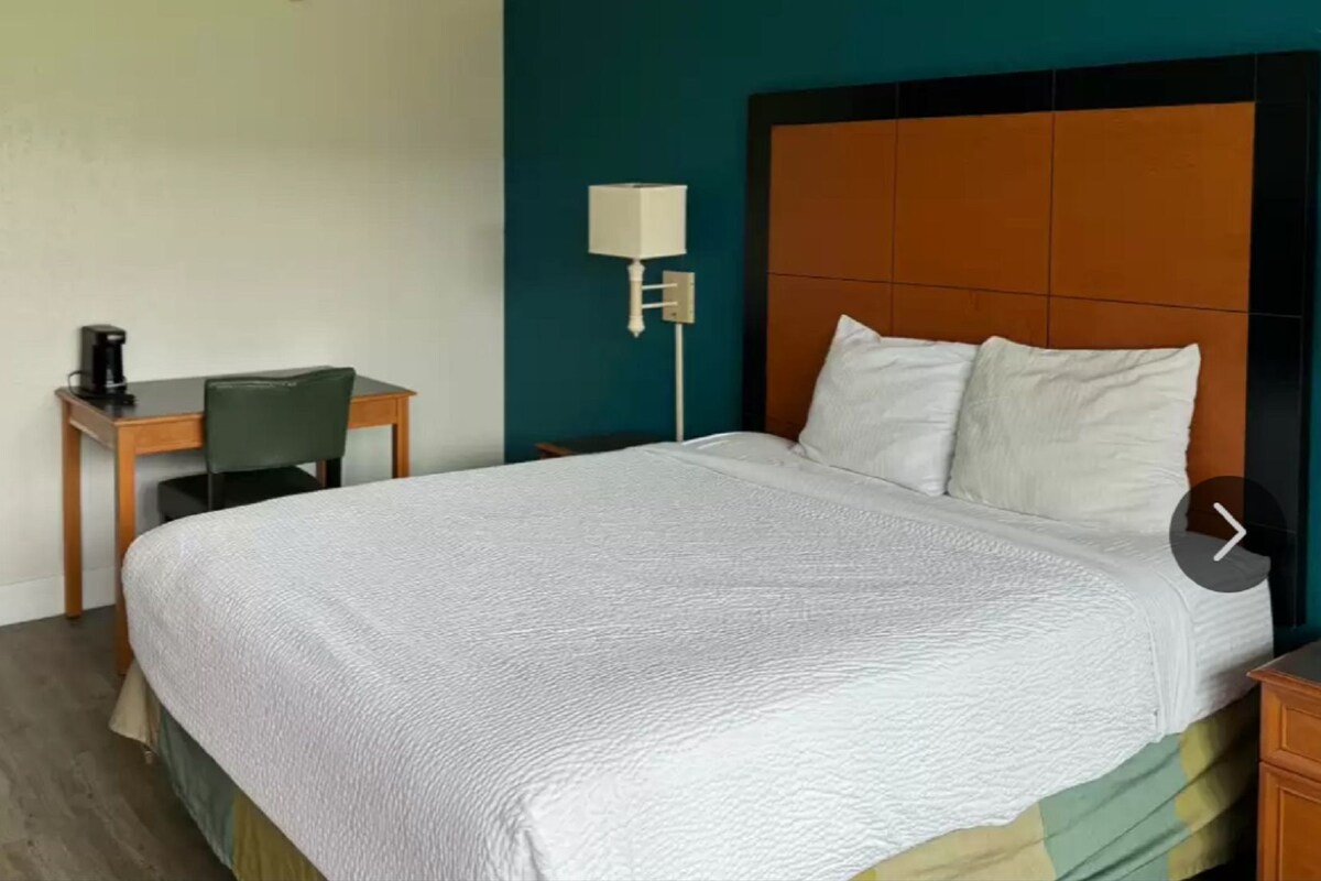 Habitación de hotel con una cama grande, un escritorio con silla, una lámpara y una cafetera.
