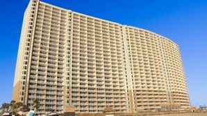 A large, multi-story hotel with a curved facade, numerous balconies, and a sandy ground level. - 2bd 2ba, Sleeps 8, Seasonal Beach Service (Panama City Beach)