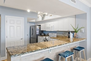 A kitchen with white cabinets, granite countertops, stainless steel appliances, and bar stools.
