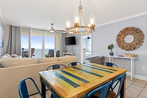 A modern living room with a large window, a dining area with a colorful table, and a chandelier.