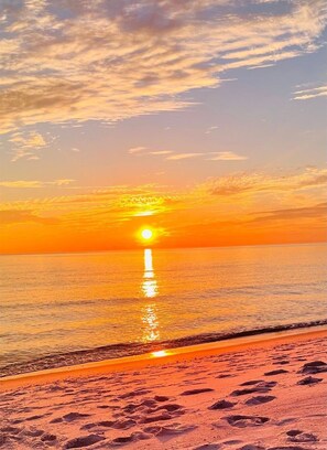 Une plage où l’on voit des empreintes de pas menant à l’eau, un coucher de soleil en arrière-plan et un ciel dégagé.