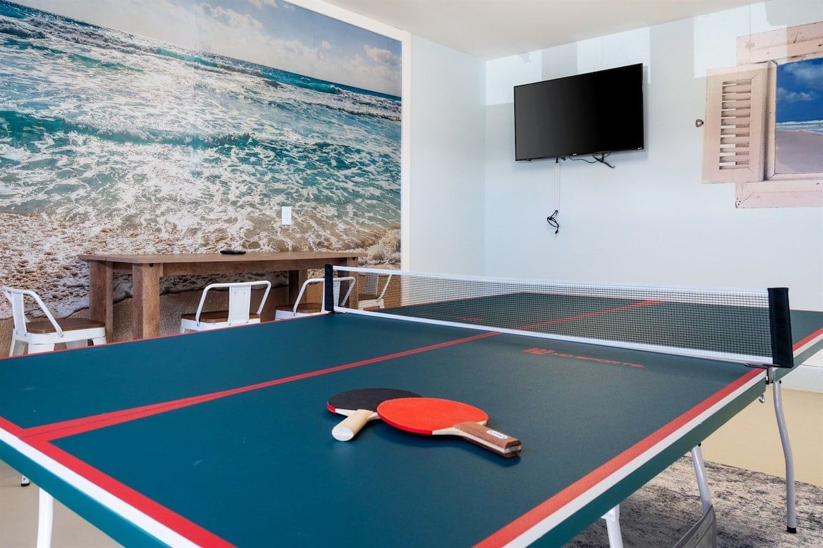 A ping pong table with two paddles, a television mounted on the wall, and a beach-themed mural.