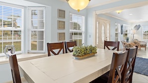 A dining area with a large table, six wooden chairs, a chandelier, and a view of the outdoors through large windows with blinds. - Sleeps 14, Game Room, Gated Beach Access, Pet Friendly (Panama City Beach)