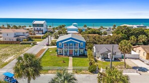 Un quartier résidentiel situé sur la côte, avec une grande maison bleue qui se distingue au centre, entourée d’autres maisons et d’une plage en arrière-plan.