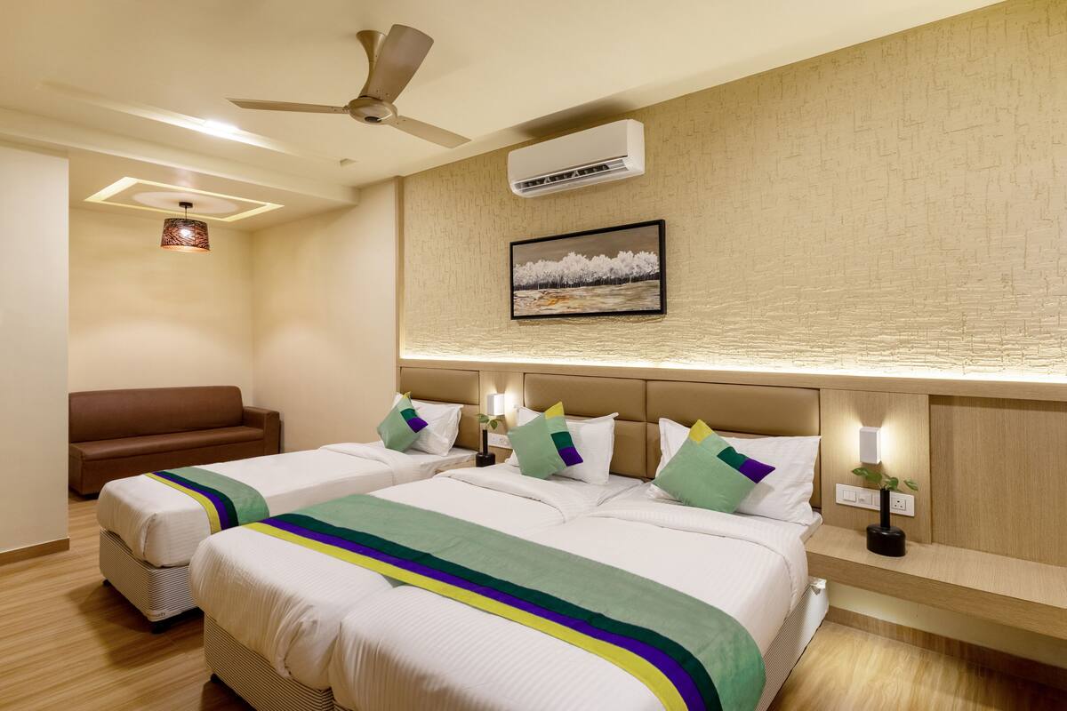 Ein Hotelzimmer mit zwei Betten, einem Sofa, einem Deckenventilator, einer Klimaanlage und einem Wandbild.