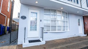 Un edificio blanco de dos pisos con puerta y ventanas blancas, una pared de ladrillo y un pequeño porche.