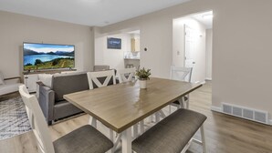 A modern living room with a wooden dining table, grey sofas, and a flat-screen TV displaying a scenic landscape. - Central Downtown Living (Buffalo)