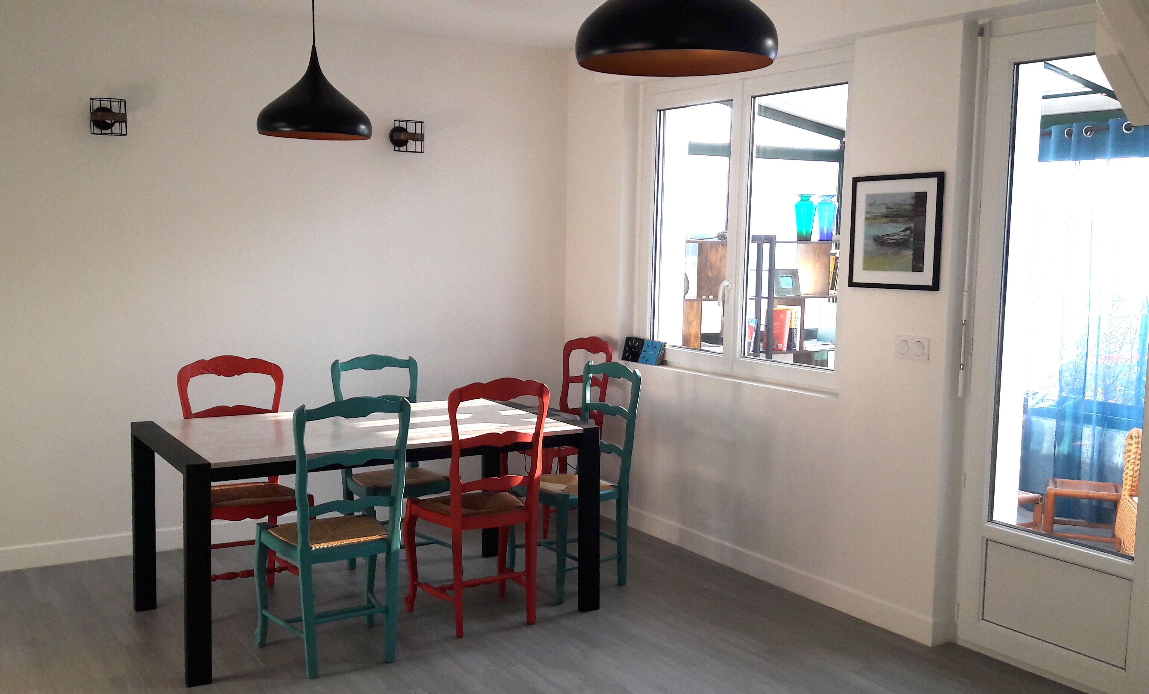 Un comedor con una mesa y sillas blancas, dos lámparas colgantes y un ventanal con vistas a muebles de exterior.