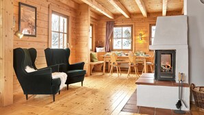 A cozy cabin interior with wooden walls, a dining area, two plush armchairs, a fireplace, and a ceiling light fixture.