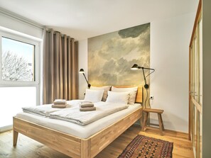 A modern bedroom with a wooden bed, two bedside lamps, a large abstract painting on the wall, and a view of trees outside the window. - The Tauplitz Suites - Alpine Suite B4 (Tauplitz)