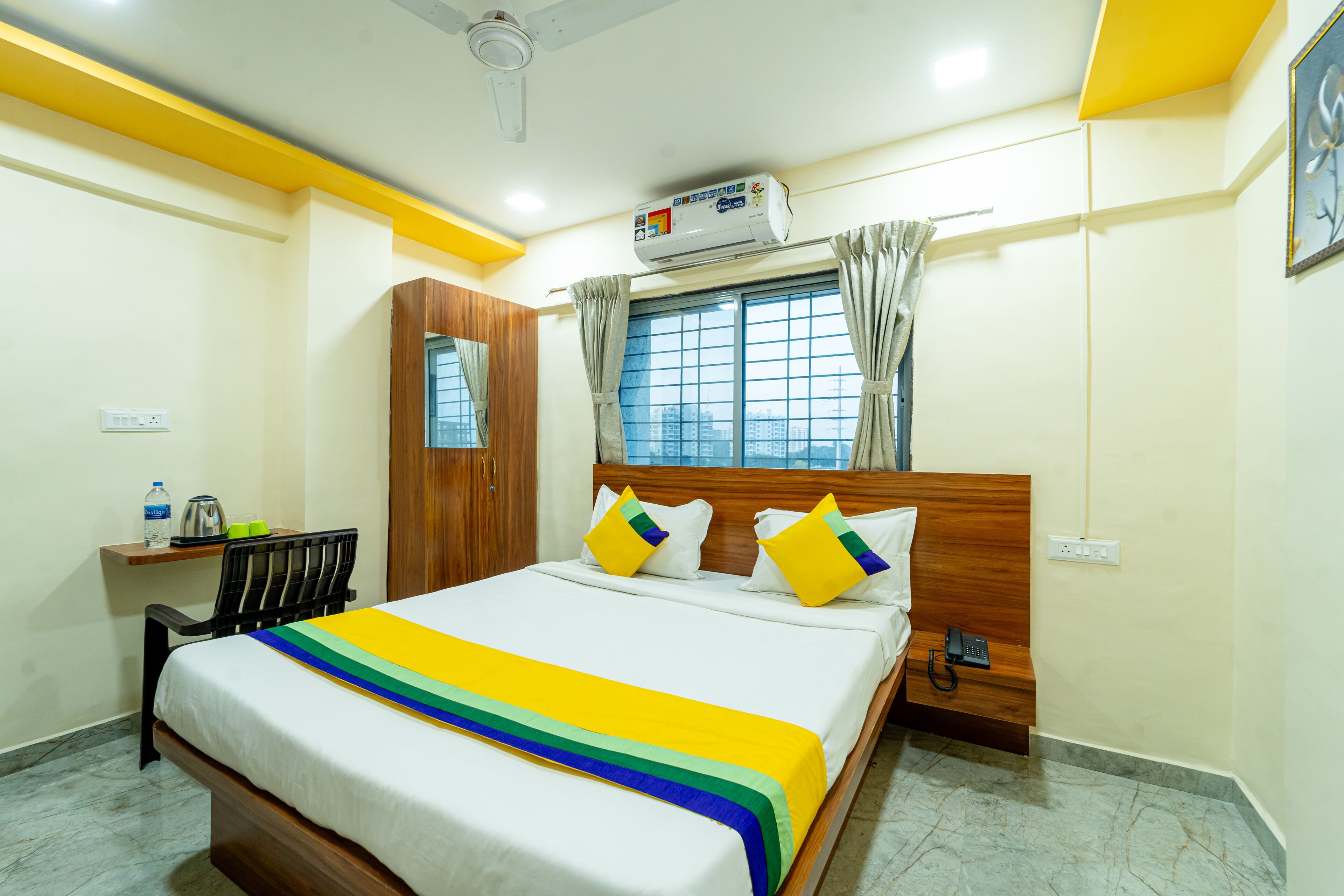 A hotel room with a bed, a wooden headboard, a window with curtains, an air conditioning unit, a bedside table with a phone, and a small shelf with a water bottle and glasses.