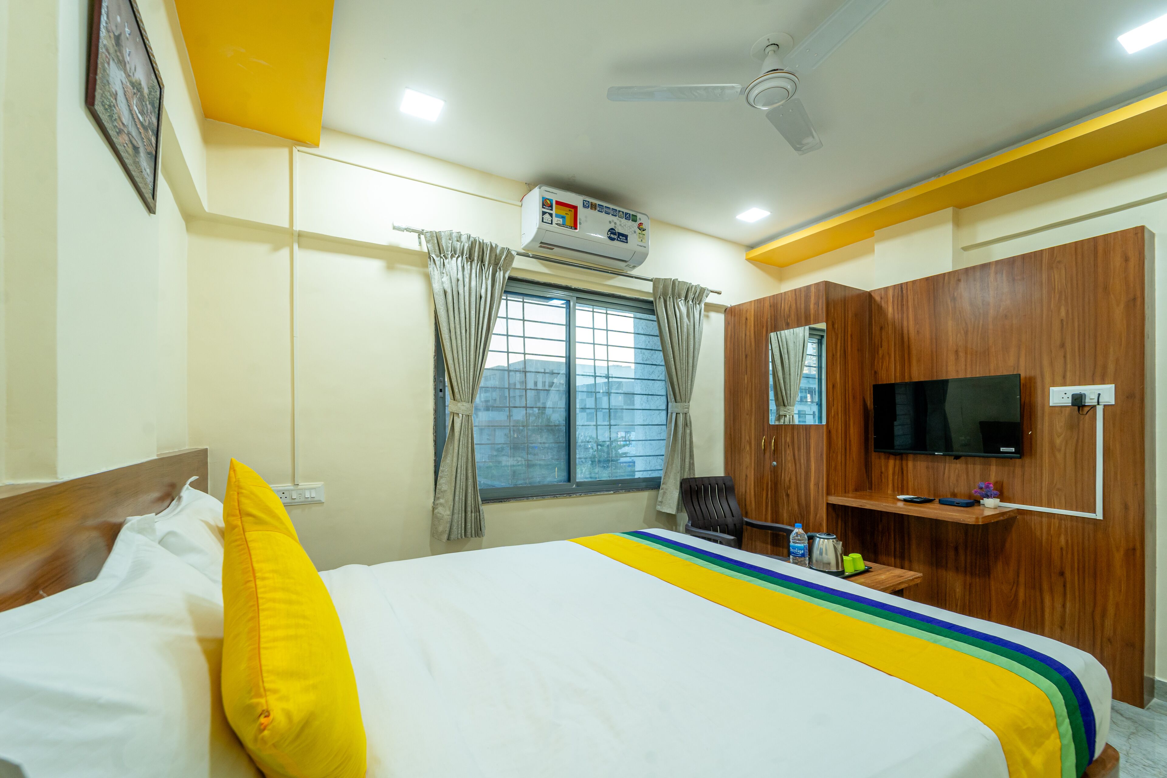 Quarto de hotel com cama, televisão, ventoinha no teto, ar condicionado, janela com cortinas, secretária com cadeira e guarda-roupa de madeira.