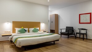 Quarto de hotel moderno com uma cama grande, mesas de cabeceira, escrivaninha, duas cadeiras e uma obra de arte emoldurada na parede.