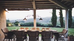 An outdoor dining area with wicker chairs and tables, a pergola, and a view of hills and trees.
