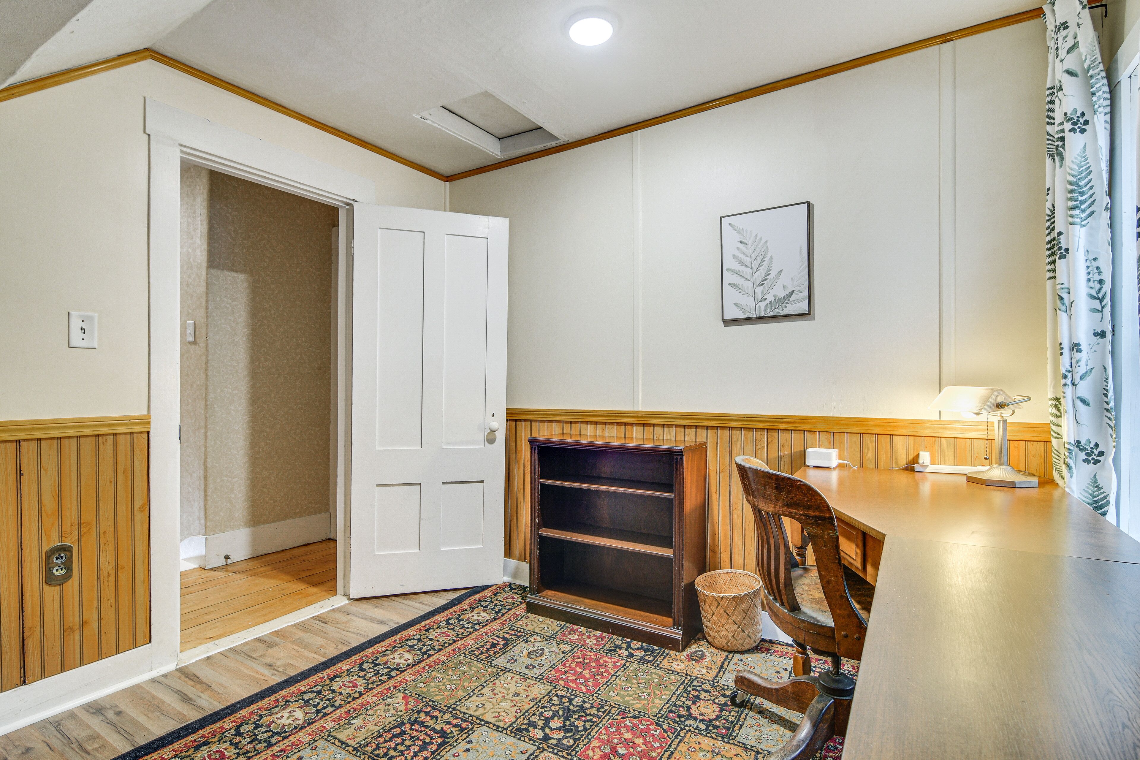 A room with a wooden floor, a patterned rug, a desk with a lamp, a chair, and a door leading to another room.