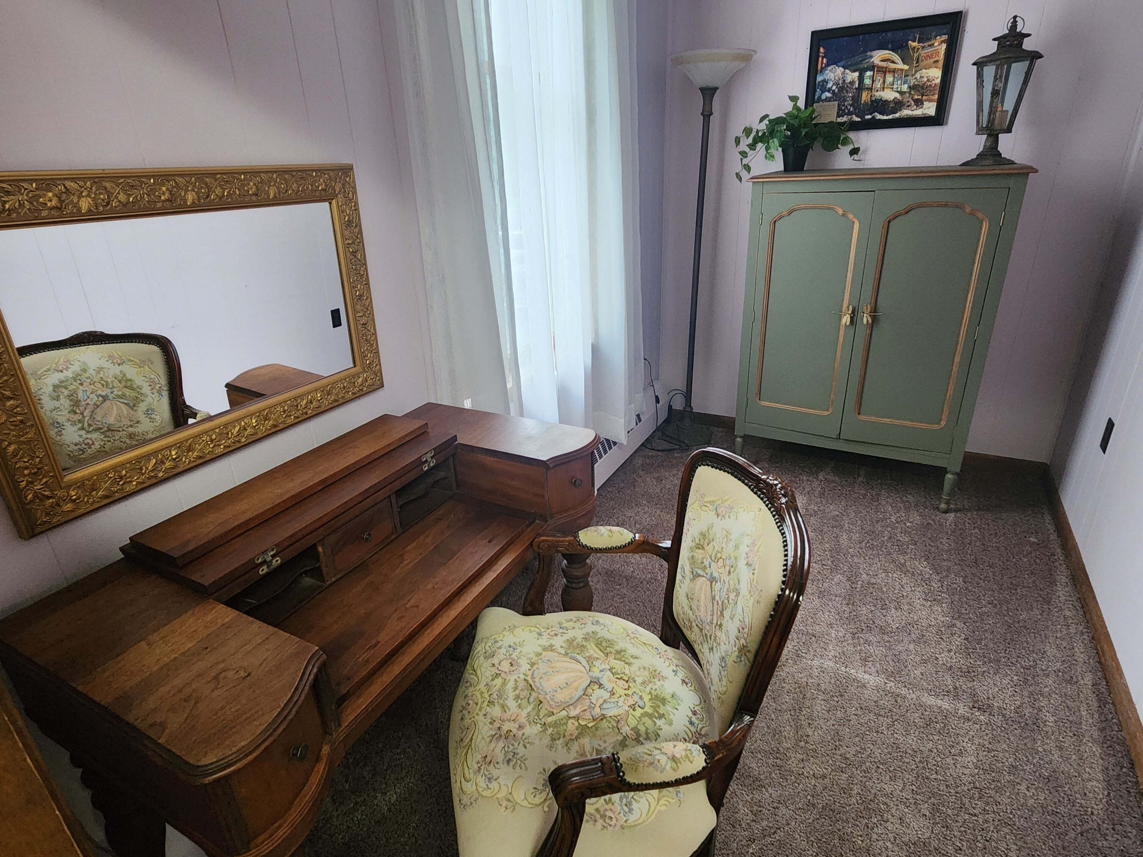 A room with a wooden desk, a chair with floral upholstery, a green cabinet, and a mirror with an ornate frame.