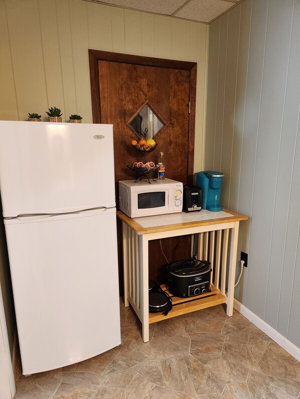 A compact kitchen with a white refrigerator, a microwave, a toaster, and a small wooden table with a slow cooker underneath.