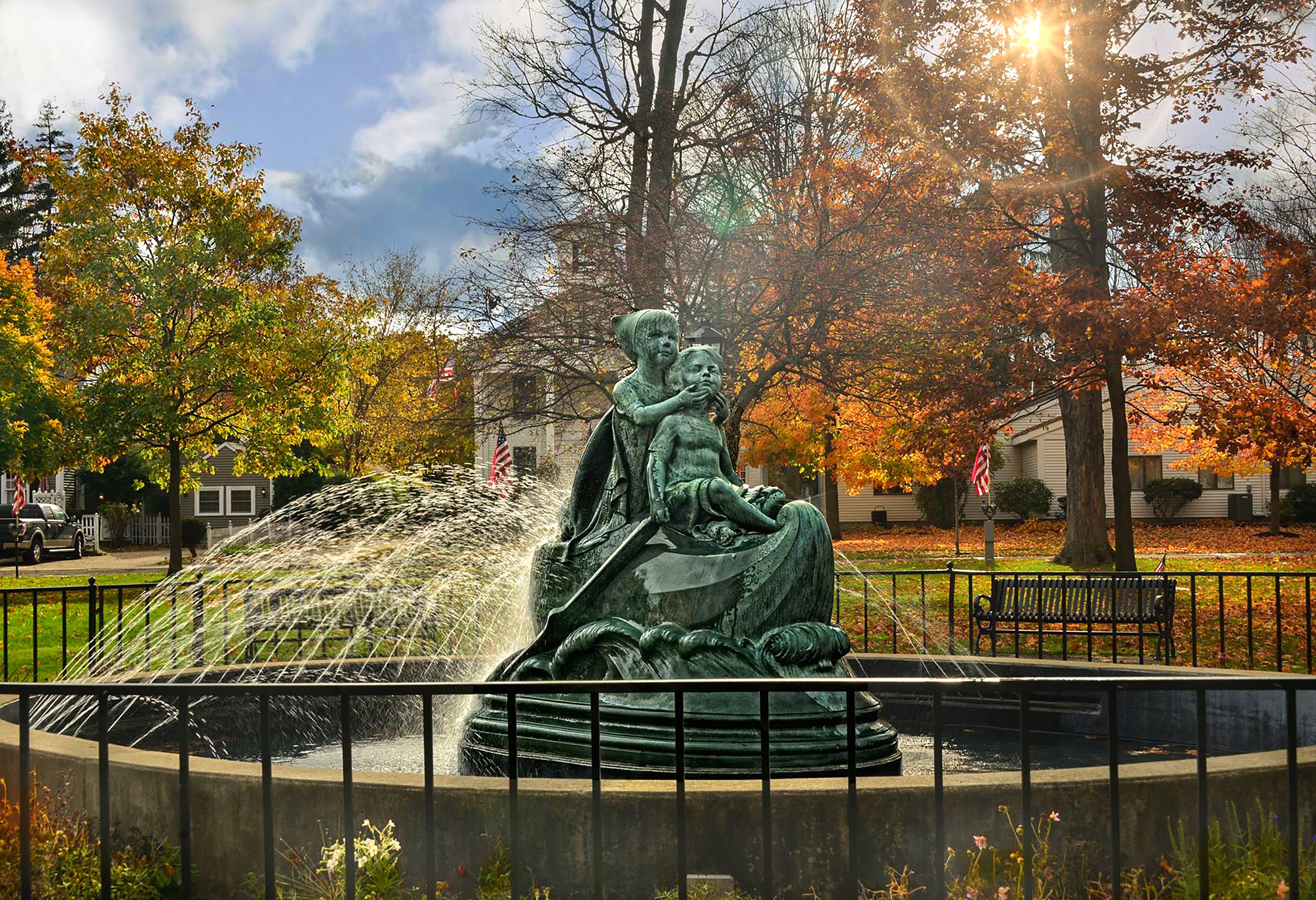 A bronze statue of a woman and child by a fountain, surrounded by autumn foliage and a bench.
