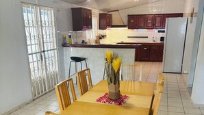 Un comedor con una mesa y sillas de madera, un florero con flores amarillas y una cocina al fondo.