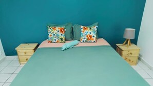 Una cama con colcha verde y dos almohadas estampadas, flanqueada por mesitas de noche de madera con lámparas, en una habitación con paredes color turquesa y suelo de baldosas blancas.