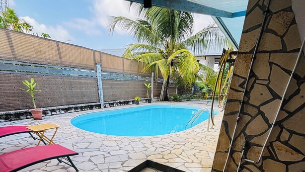 Zona de piscina con baldosas de piedra, una silla de descanso roja y una silla amarilla, rodeada de palmeras y una valla de metal.
