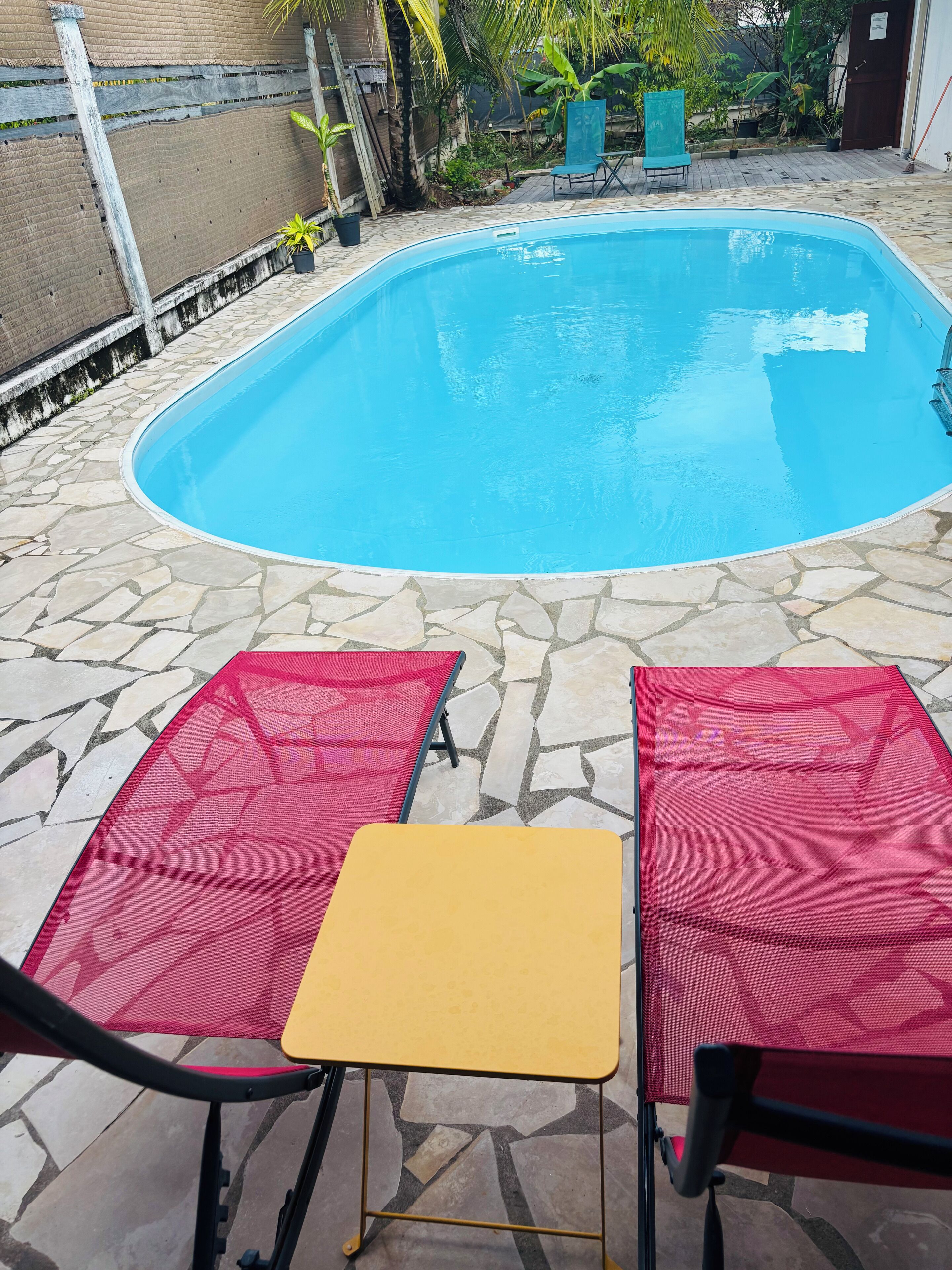 A swimming pool with a stone tile deck, two red and one yellow lounge chair, and a small table.