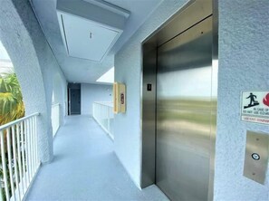 Un couloir avec un mur blanc, un ascenseur en métal et une porte au fond.