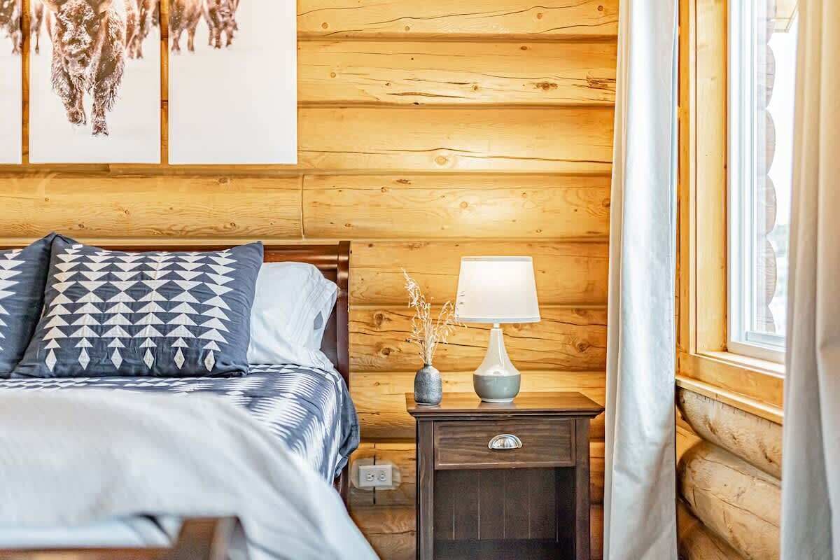 Ein Schlafzimmer mit einer Wand aus Holzbalken, einem Bett mit gemusterten Kissen, einem Nachttisch mit Lampe und einem Fenster mit Vorhängen.