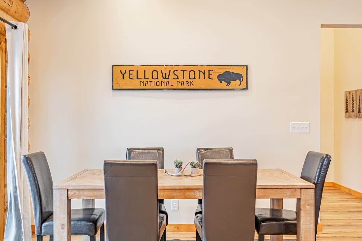 Uma área de refeições com uma mesa de madeira e quatro cadeiras, um letreiro fixado na parede com a inscrição "Yellowstone National Park" e um pequeno vaso de planta sobre a mesa.