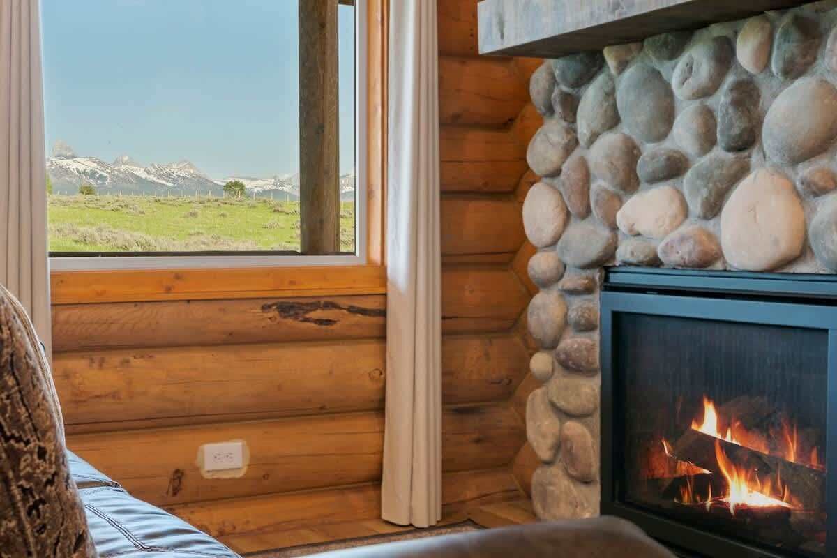 A log cabin interior with a stone fireplace, a window with a view of a green field and mountains, and a cozy seating area.
