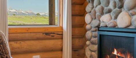A log cabin interior with a stone fireplace, a window with a view of a green field and mountains, and a cozy seating area.