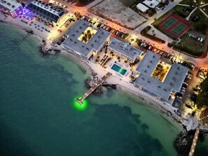 An aerial view of a coastal hotel with a pool, tennis courts, and a pier extending into the water.