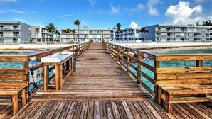 A wooden pier leading to a beachfront hotel with balconies, palm trees, and a clear sky. - Salty Breezes Condo 2bed 2bath second floor with elevator, pool & ocean front! (Key Colony Beach)
