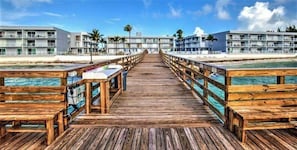 A wooden pier leading to a beachfront hotel with balconies, palm trees, and a clear sky.