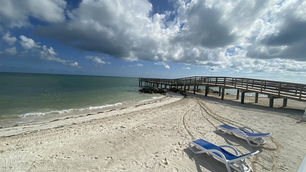 A beach with a wooden pier extending into the water, clear skies, and footprints in the sand. - Salty Breezes Condo 2bed 2bath second floor with elevator, pool & ocean front! (Key Colony Beach)