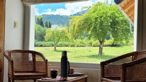 Une pièce avec deux fauteuils en osier, une petite table avec une bouilloire, et une vue sur un paysage verdoyant par une grande fenêtre.