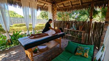 Experience a serene spa retreat with a massage table nestled under a thatched roof, surrounded by lush greenery.